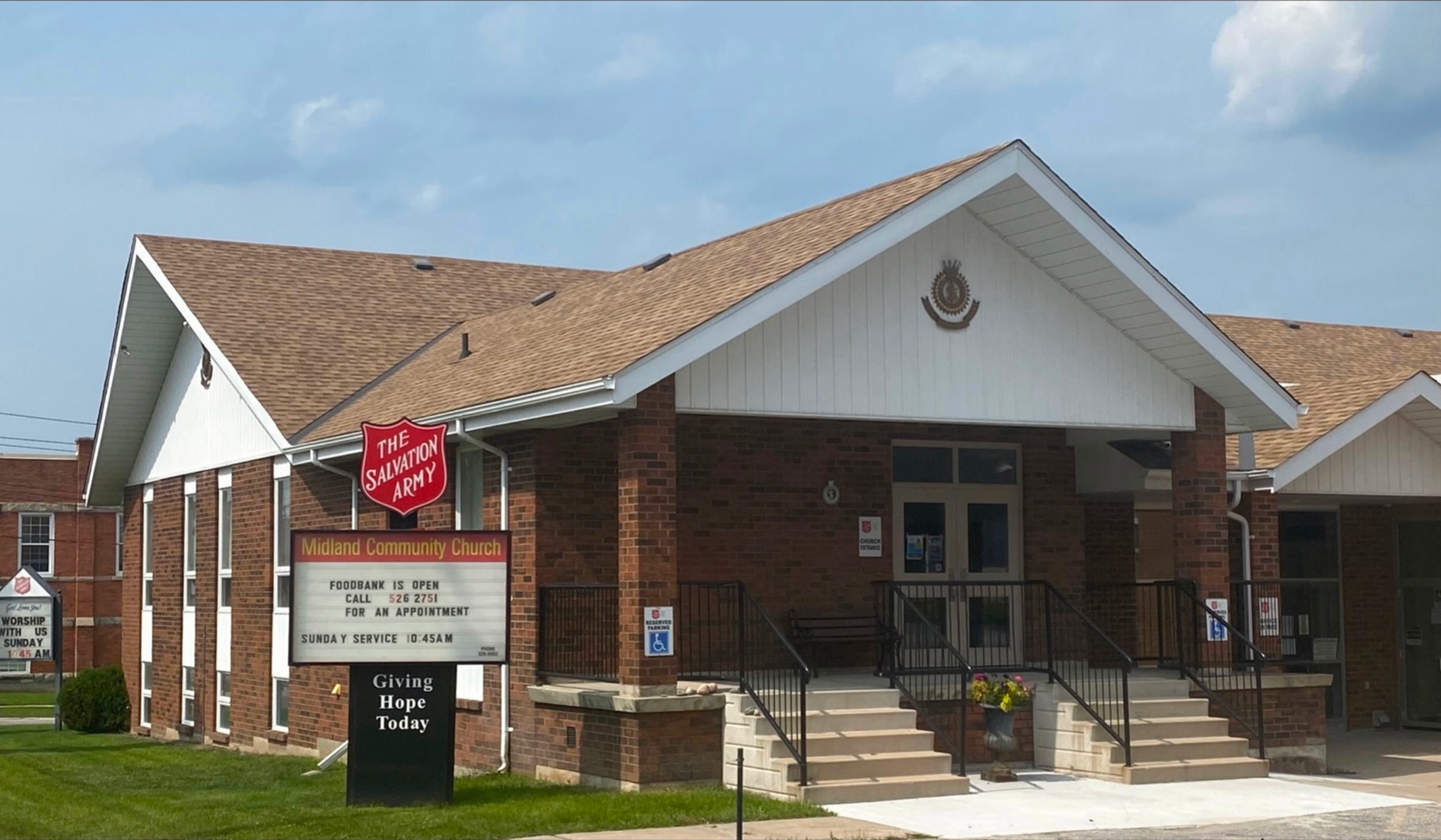 Exterior of the Midland Salvation Army Community Church, a brick building with a peaked roof, accessible entrance, and signboard displaying foodbank and Sunday service information.