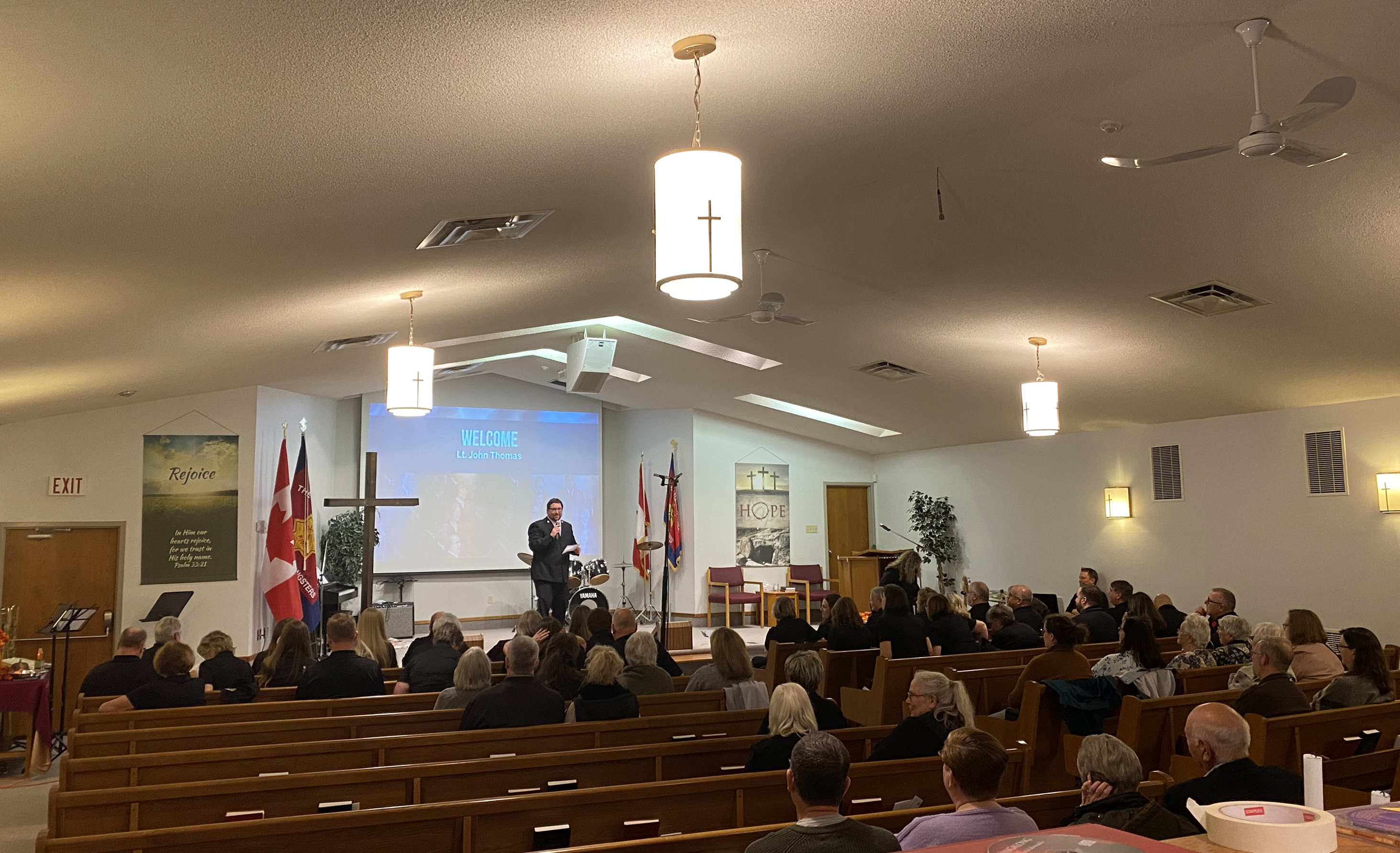 Interior of the Midland Salvation Army church sanctuary during a service, with a pastor speaking at the front near Canadian and Salvation Army flags while the congregation sits in wooden pews.