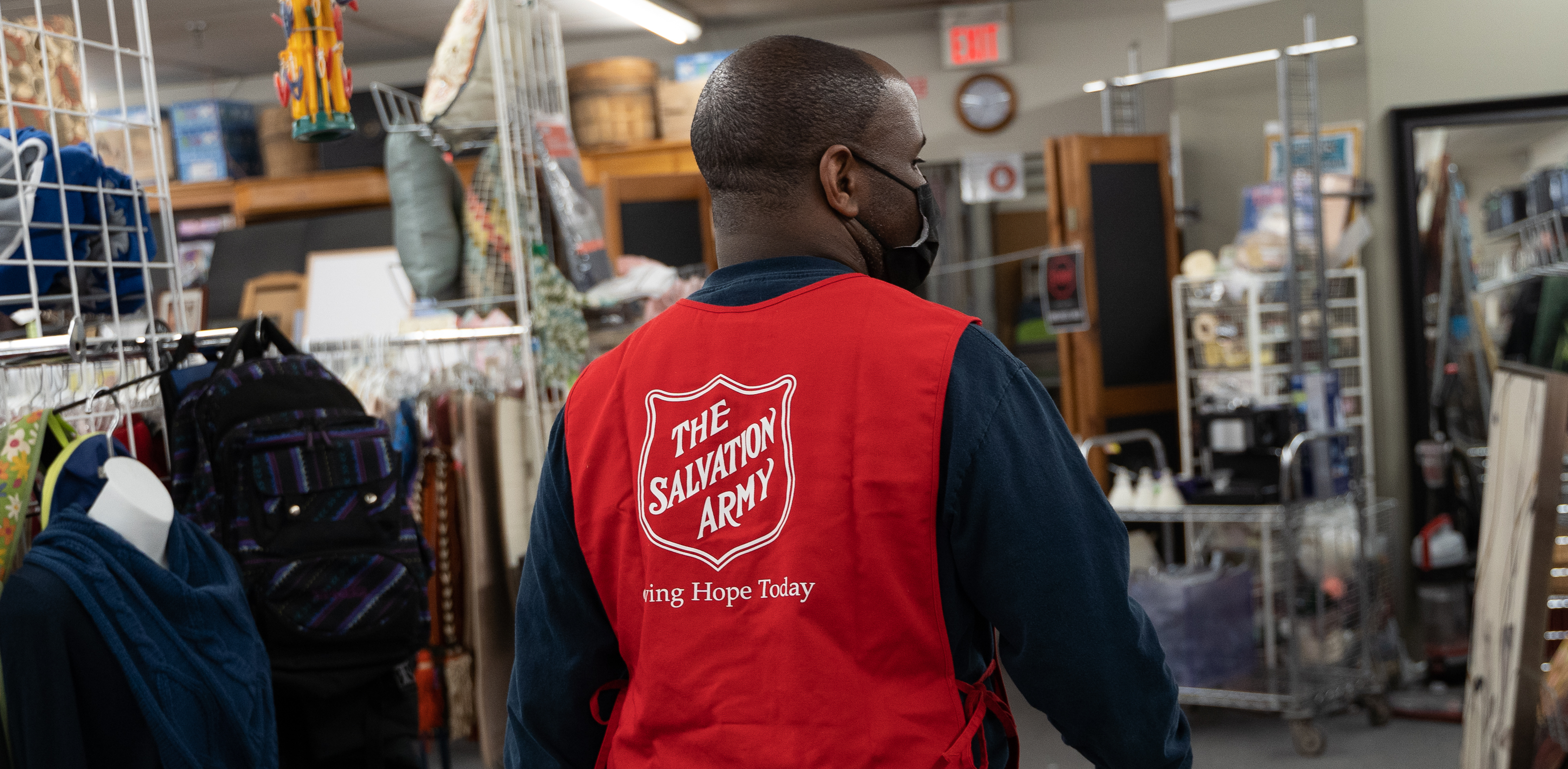 Person wearing a red Salvation Army vest walking through a thrift store aisle with clothing racks and items on display.
