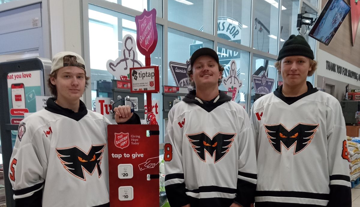 Three people in hockey jerseys standing beside a Salvation Army donation kettle and sign inside a store.