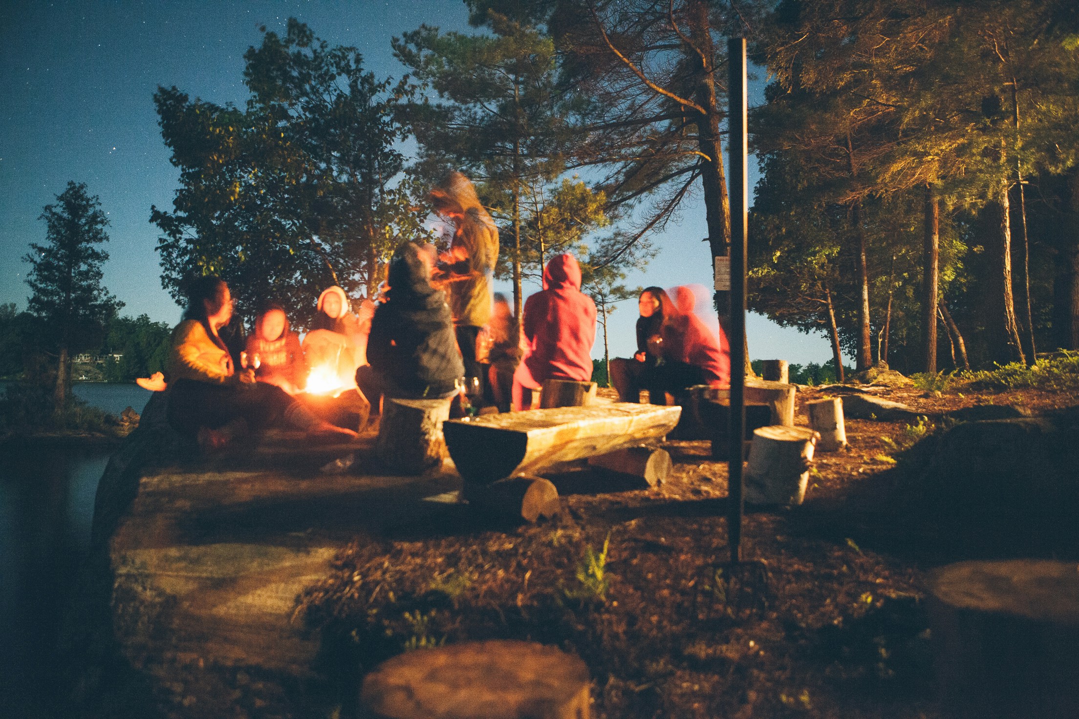 Group of people sitting and standing around a campfire at night in a wooded area, with logs used as benches and a lake visible in the background.