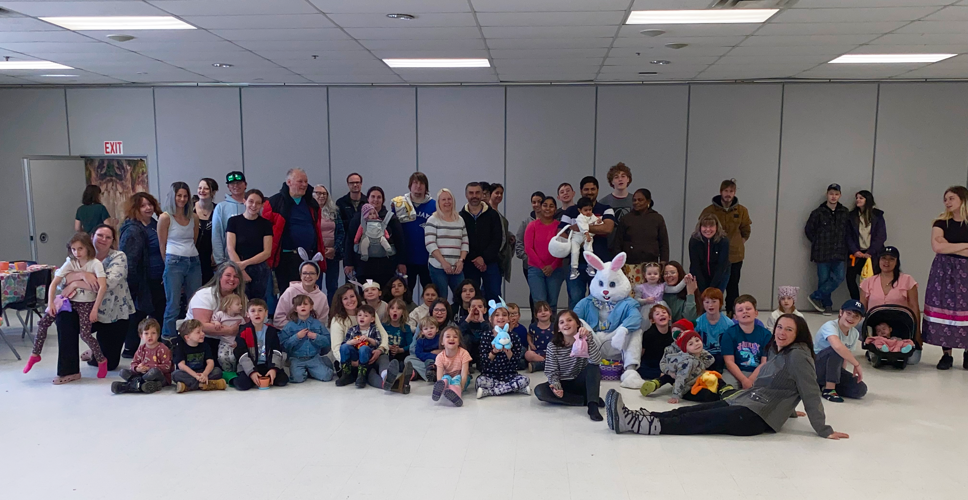 Large group of adults and children gathered indoors, posing together in a community hall, with some children seated on the floor and others standing behind.