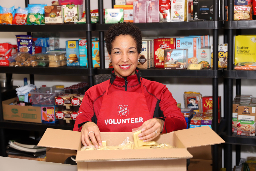 Smiling volunteer wearing a Salvation Army apron packs food items into a cardboard box, standing in front of fully stocked shelves with boxed and canned goods in a food pantry setting.