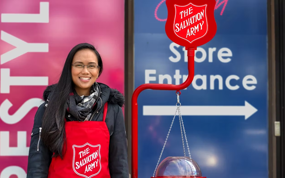 Smiling volunteer wearing a red Salvation Army apron standing beside a donation kettle and sign outside a store entrance.